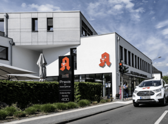 Außenansicht der Kloster Apotheke in einem modernen weißen Gebäude mit großem Apothekenzeichen und Straßenschild